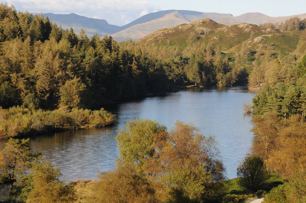 A wide gravel path curving around the shore of Tarn Hows with fells reflected in still water, a classic easy Lake District walk